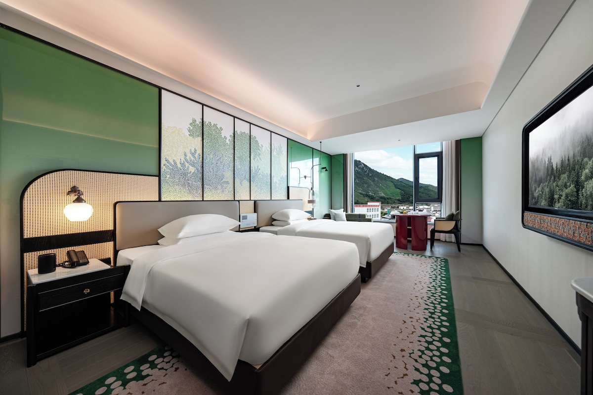 A modern hotel room with two beds, a large window, and a view of a mountainous landscape.