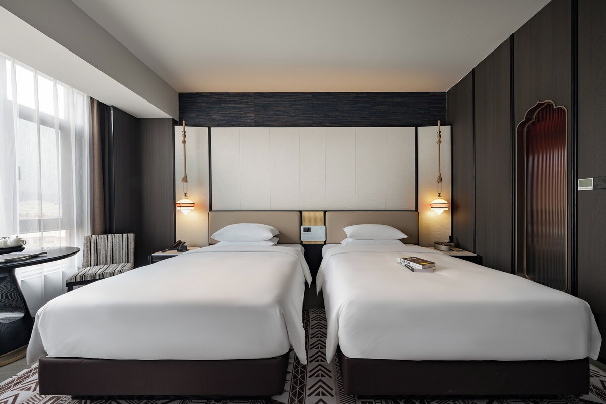A hotel room with two beds, a desk, a chair, and a large window.