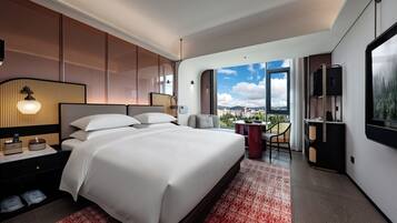 A modern hotel room with a large bed, a desk, a chair, a TV, and a balcony with a city view.