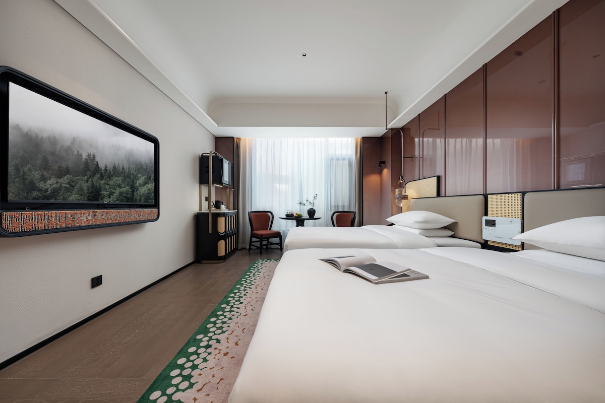 A hotel room with a large bed, a desk, a chair, a television, and a view of a forest through the window.