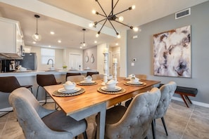 Una cocina moderna con zona de comedor, que incluye una mesa de madera preparada para cuatro personas, sillas tapizadas en gris y una lámpara colgante contemporánea.