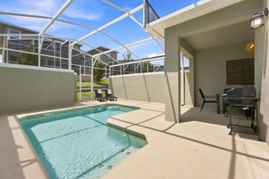Un área de piscina con cerramiento de vidrio, una parrilla y mobiliario exterior.