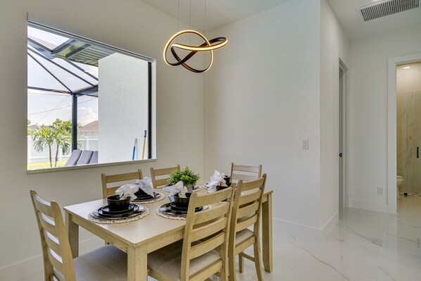 A dining area with a table set for four, a modern pendant light, and a view through a large window. - Private Pool & Fun-Filled 4BR Cape Coral Retreat (Cape Coral)