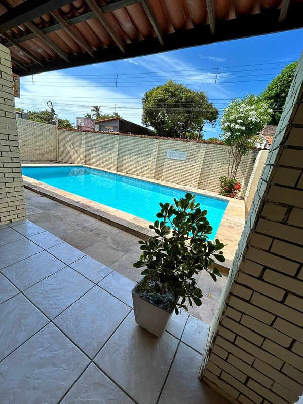A swimming pool with a tiled deck, surrounded by a brick wall and a covered area with a metal roof.