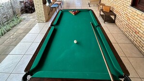 Una mesa de billar con taco, rodeada de sillas y mesas, bajo una zona techada con pared de ladrillo y techo de madera.