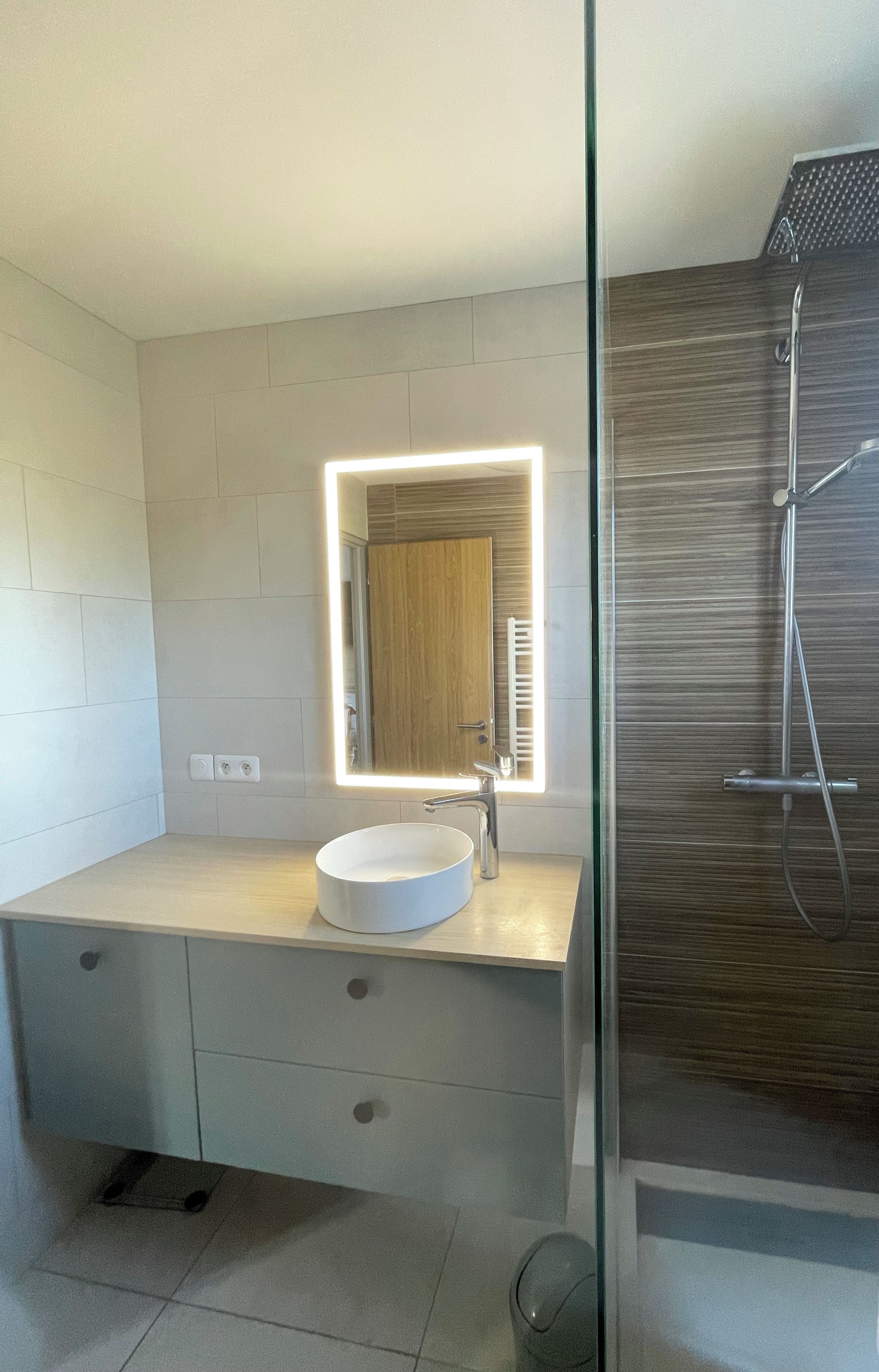 Une salle de bain moderne avec un lavabo blanc, un miroir avec éclairage intégré et une douche.