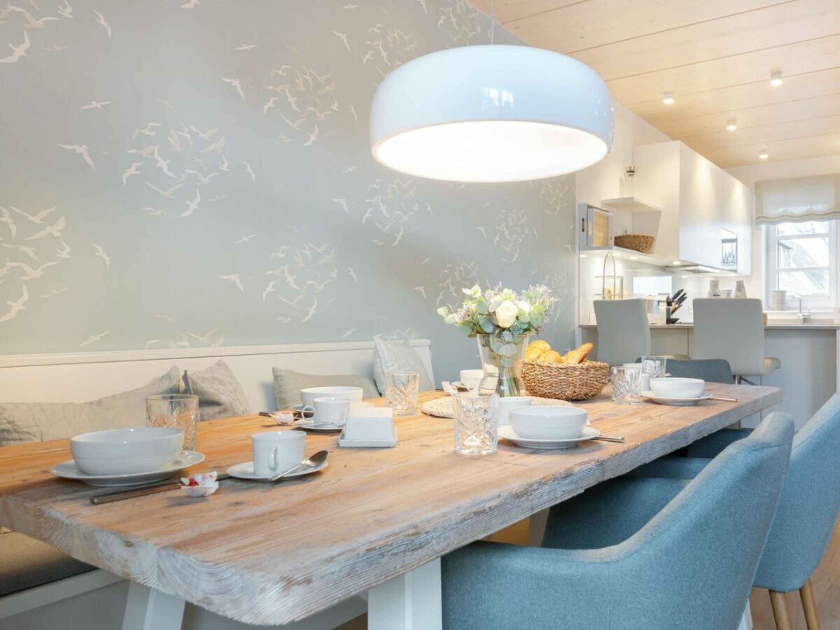 A dining area with a wooden table set for two, a vase with flowers, and a hanging light fixture.