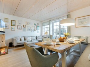 A cozy living room with a sofa, a dining table set for two, and a large window providing natural light. - Holiday Home Watt'n Gluck - New! (Sylt)