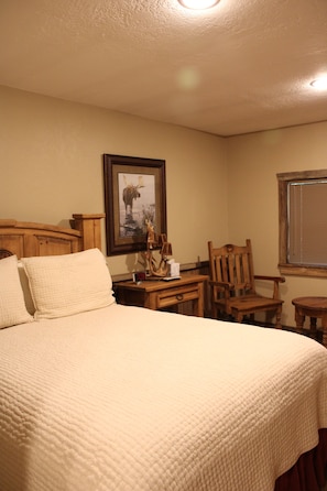 A hotel room with a bed, a wooden nightstand, a framed picture of a moose, a chair, and a window with blinds. - Private East Texas Ranch Retreat with 7 Cabins, Trophy Fishing & More (San Augustine)