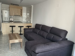 Et moderne opholdsrum med en mørk lædersofa, et træbord og et tekøkken med køleskab, vask og barstole.