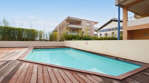 A rectangular swimming pool with a wooden deck surrounding it, set against a backdrop of buildings and greenery. - Apartment with balcony and swimming pool (Grasse)