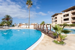 A swimming pool with a wooden deck, palm trees, and a building with balconies. - Vrbo Property (Adeje)