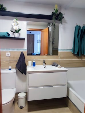 A bathroom with a toilet, sink, and bathtub. There is a towel rack with towels, a plant on the wall, and a mirror reflecting the bathroom. - Vrbo Property (Adeje)