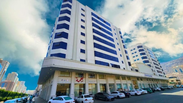 Et hotel i flere etager med en moderne facade, en indgang i stueplan og en parkeringsplads med flere biler.