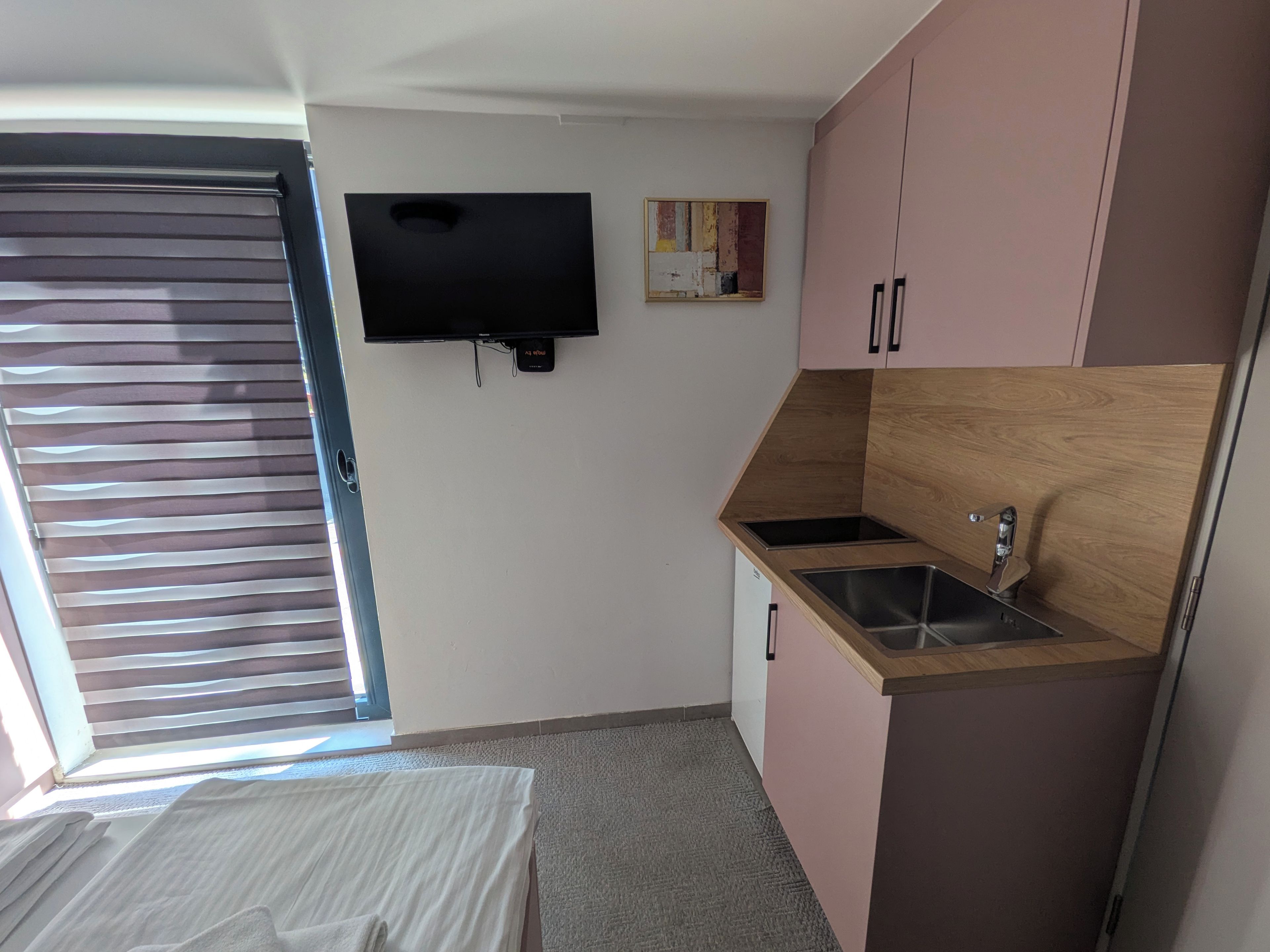 Une pièce de taille modeste comprenant un lit, une petite kitchenette, un téléviseur fixé au mur et une fenêtre avec des stores.