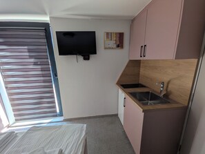 A compact room with a bed, a small kitchenette, a TV mounted on the wall, and a window with blinds.