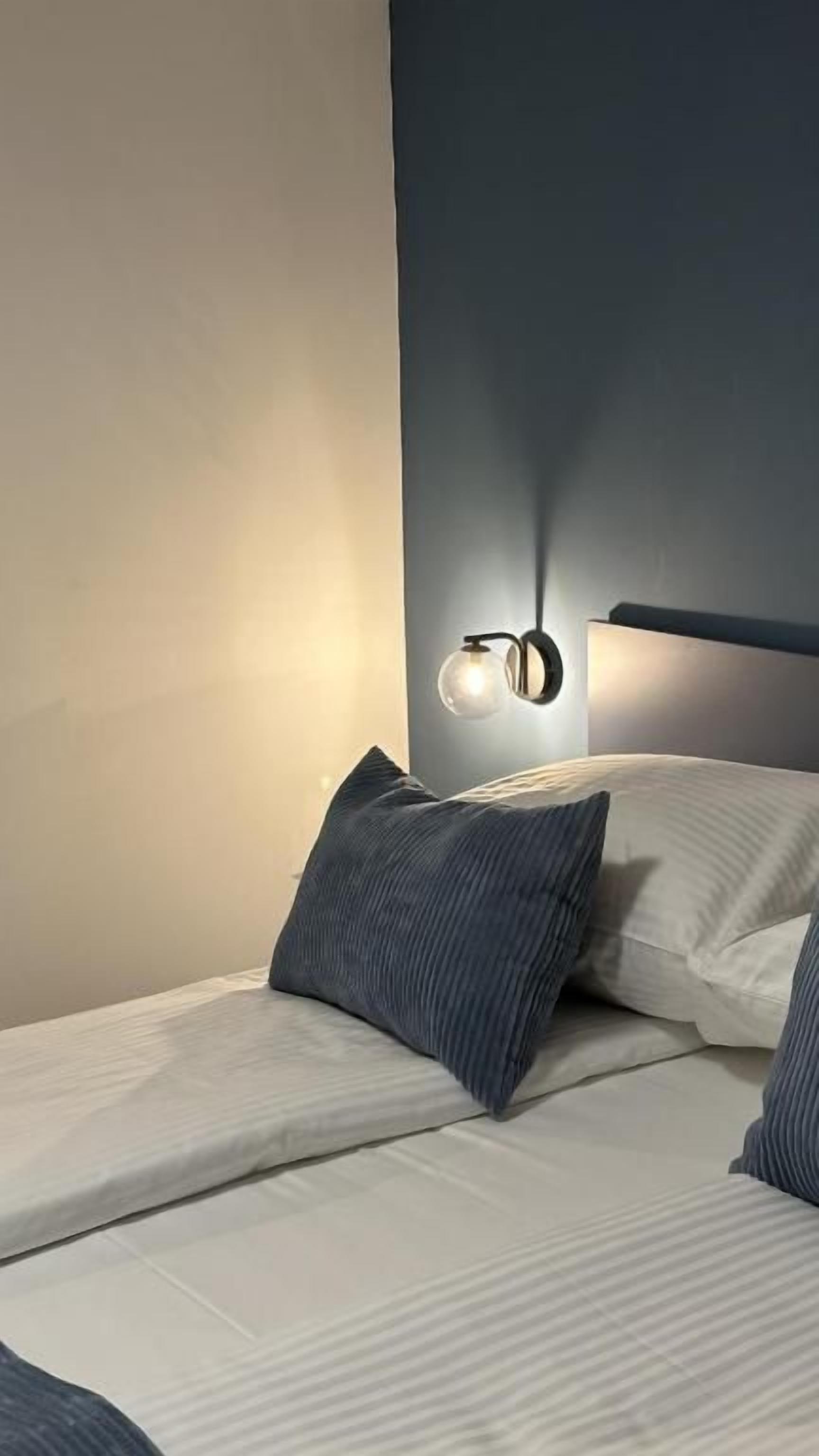 Una cama con cabecera, una lámpara de pared y un almohadón.