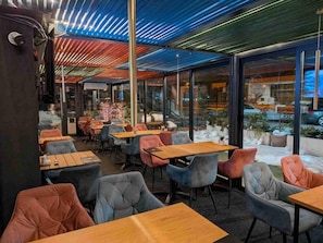 An indoor seating area with tables and chairs, large windows, and a ceiling with a patterned design. - Alinar Oasis Budget Hotel (Sarajevo)