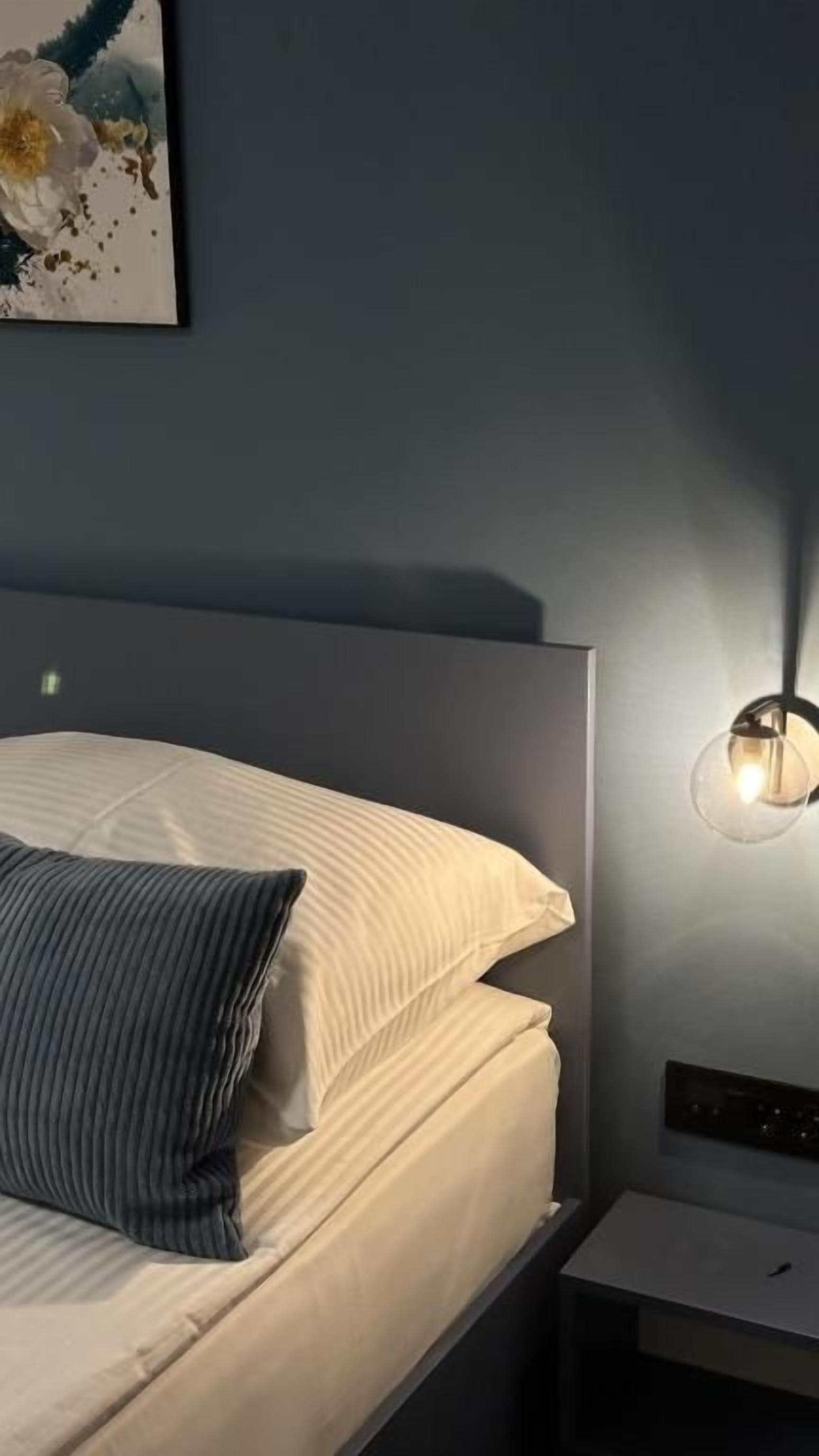 Una cama con ropa de cama blanca y una almohada a rayas, una mesita de noche y una lámpara de pared.