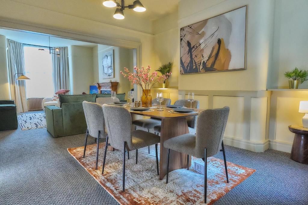 A dining area with a round table, six chairs, and a vase with flowers. There is a painting on the wall and a plant on a side table.