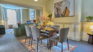 A dining area with a round table, six chairs, and a vase with flowers. There is a painting on the wall and a plant on a side table.