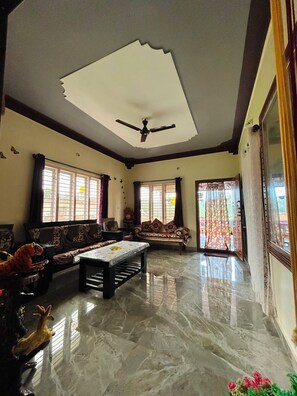 A spacious living room with a ceiling fan, a sofa set, a coffee table, and a large window with curtains.