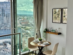 A dining area with a view of a cityscape through a large window, a round table with chairs, and a wooden shelf with decorative items. - Skyline Rotunda Studio Apartment (Birmingham)