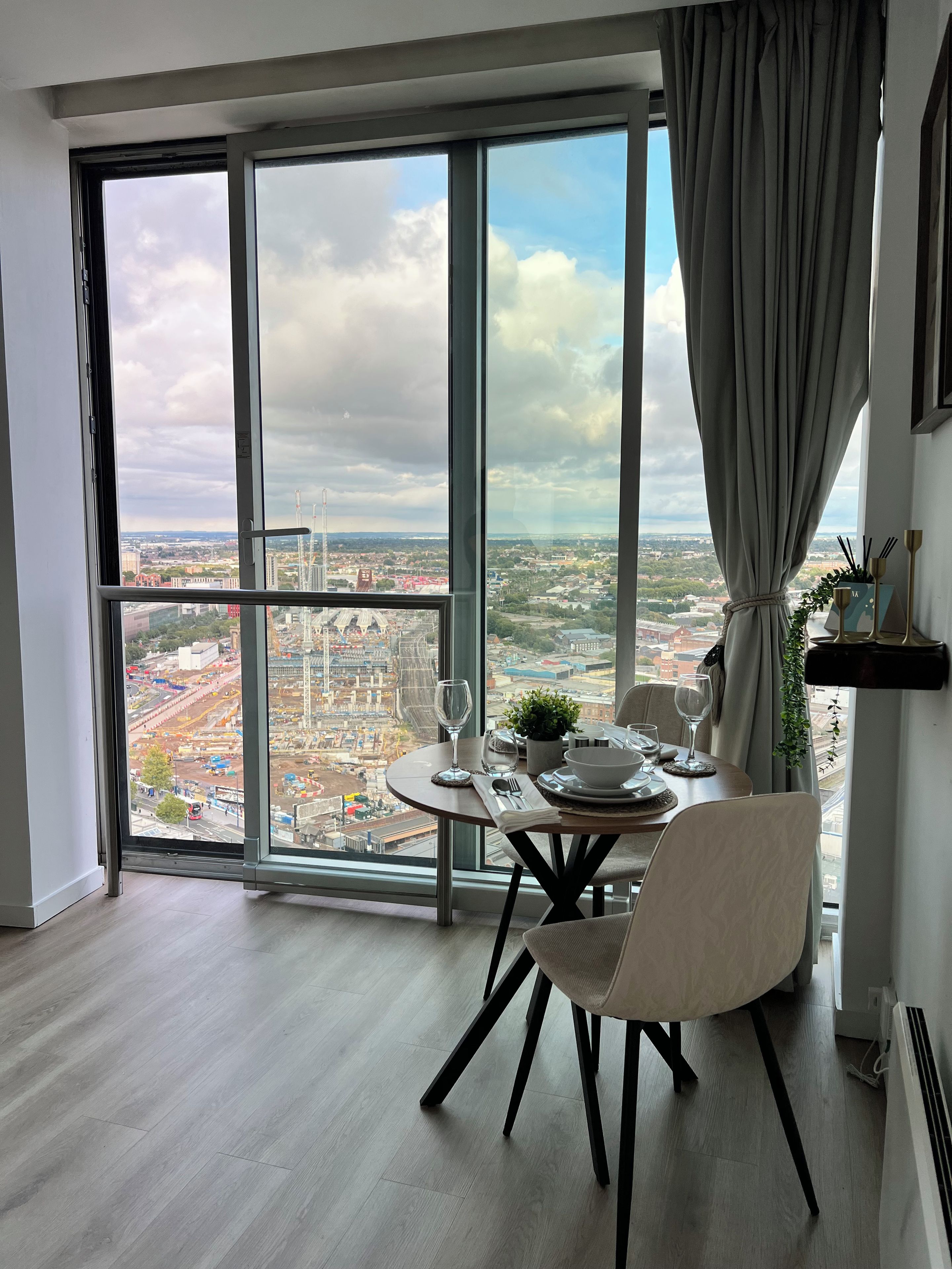 Un coin repas avec une table dressée pour deux personnes, une vue sur le paysage urbain à travers de grandes fenêtres et un rideau à moitié tiré.