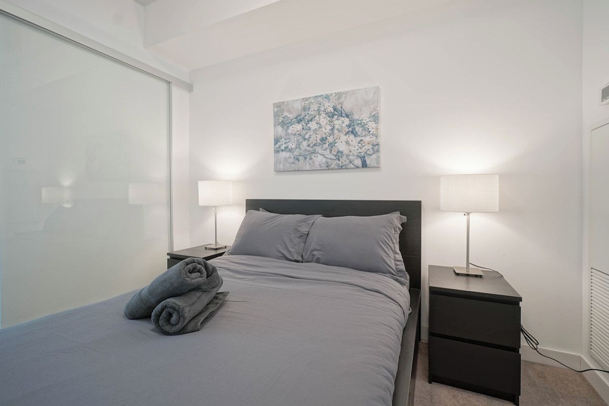 A bedroom with a bed, two bedside tables, a wall-mounted lamp, and a painting.