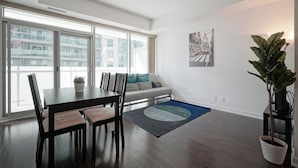 A modern living room with a dining table, chairs, a sofa, a large window, and a potted plant. - Bright 1Bedroom w/ Balcony Steps to CN Tower-MTCC (Toronto)