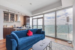 A modern living room with a blue sofa, a dining table, and a large window offering a city view. - Cozy Toronto Condo with CN Tower View & Workspace (Toronto)