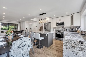 A modern kitchen with a central island, stainless steel appliances, and a dining area with a large window. - 5BR North York Home - Sleeps 11 - Parking (Toronto)