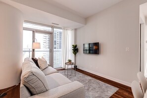 A modern living room with a sofa, a TV mounted on the wall, and a view of the city through large windows. - 2BR Condo w/ Stunning CN Tower View & Free Parking (Toronto)