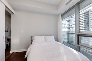 A bedroom with a bed, a window with a view of buildings, and a door leading to another room. - 2BR Condo w/ Stunning CN Tower View & Free Parking (Toronto)