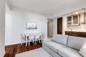 A modern living room with a grey sofa, a dining table with chairs, and a kitchen area with cabinets and a countertop. - 2BR Condo w/ Stunning CN Tower View & Free Parking (Toronto)