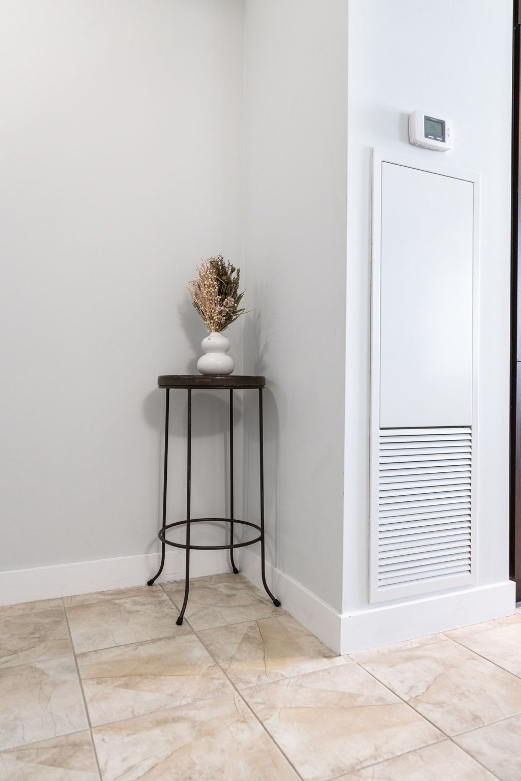A corner of a room with a white wall, a white door with a vent, and a small black metal table with a vase of dried flowers.