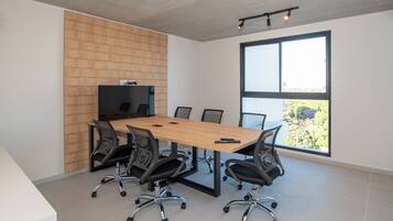 A conference room with a long table, six chairs, a television on a wooden wall, and a large window with a view of greenery.