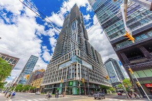 A modern high-rise building with a prominent corner entrance, surrounded by other tall buildings and a clear sky. - Downtown Toronto Studio with City View Balcony (Toronto)