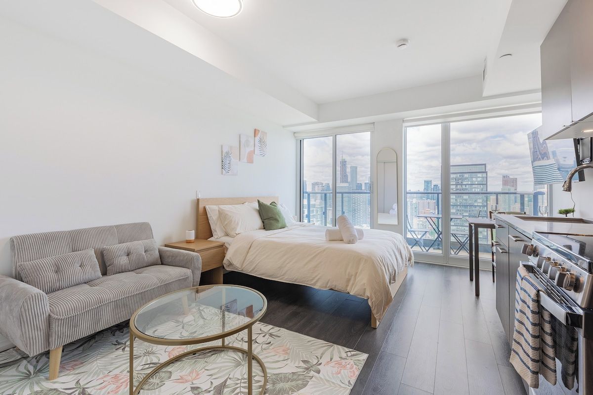 A modern apartment with a large bed, a sofa, a glass coffee table, a kitchen area, and a city view.