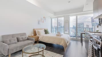 A modern apartment with a large bed, a sofa, a glass coffee table, a kitchen area, and a city view.