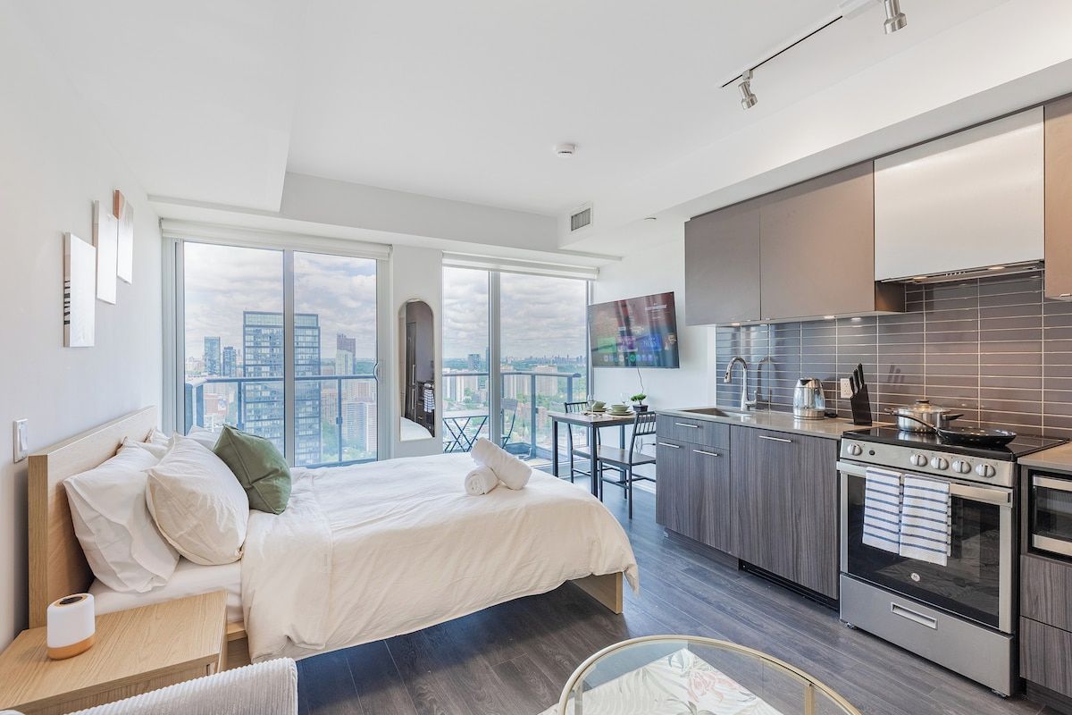 A modern hotel room with a large bed, a kitchenette, and a city view.