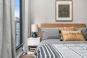 A bedroom with a bed, a nightstand, a lamp, a framed artwork, and a window with curtains. - Modern Downtown Studio with Balcony & City Views (Toronto)