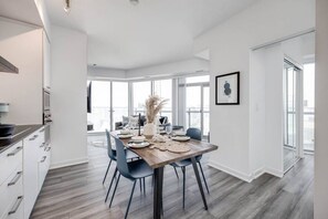 A modern dining area with a wooden table set for four, surrounded by chairs, in a room with large windows and a view of the city. - 2BRAPT w/ Parking&WFH - CN tower,MTCC,Scotia Arena (Toronto)