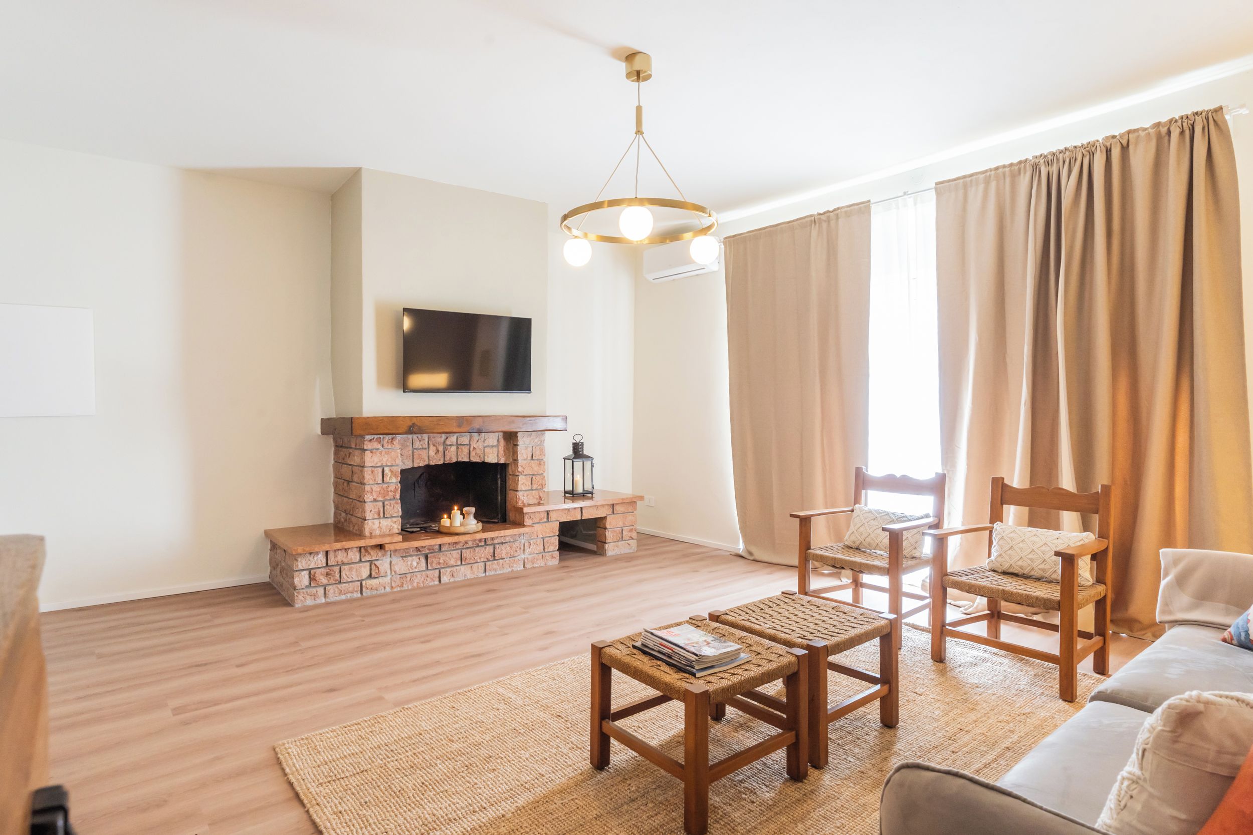 Ein Wohnzimmer mit Kamin aus Ziegelsteinen, einem flachbildfernseher, der darüber montiert ist, und einer modernen Kristallleuchter. Zwei Holzstühle und ein kleiner Tisch mit Zeitschriften stehen dort. Die Wände sind in hellen Farben gehalten und ein großes Fenster mit beigen Vorhängen sorgt für Licht.