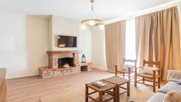 Ein Wohnzimmer mit Kamin aus Ziegelsteinen, einem flachbildfernseher, der darüber montiert ist, und einer modernen Kristallleuchter. Zwei Holzstühle und ein kleiner Tisch mit Zeitschriften stehen dort. Die Wände sind in hellen Farben gehalten und ein großes Fenster mit beigen Vorhängen sorgt für Licht.