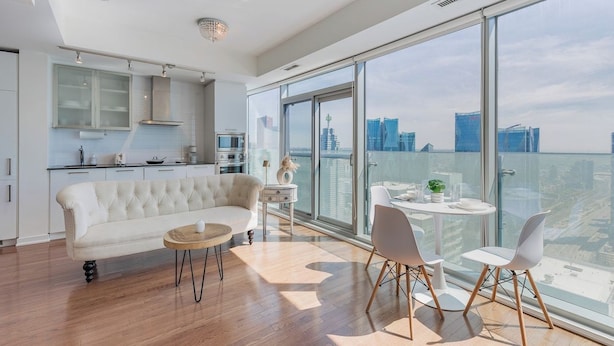 Een moderne woonkamer met een bank, een ronde tafel, stoelen, een keukenhoek en grote ramen van de vloer tot aan het plafond met uitzicht op de stad.