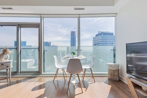 Ein modernes Wohnzimmer mit einer großen Schiebeglasdoor, einem Essbereich mit weißen Stühlen, einem kleinen Tisch und einem Flachbildfernseher.