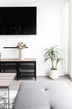 Ein modernes Wohnzimmer mit einem an der Wand befestigten Flachbildfernseher, einem Holztischchen mit Blumen in einer Vase, einer Topfpflanze und einem grauen Sofa.