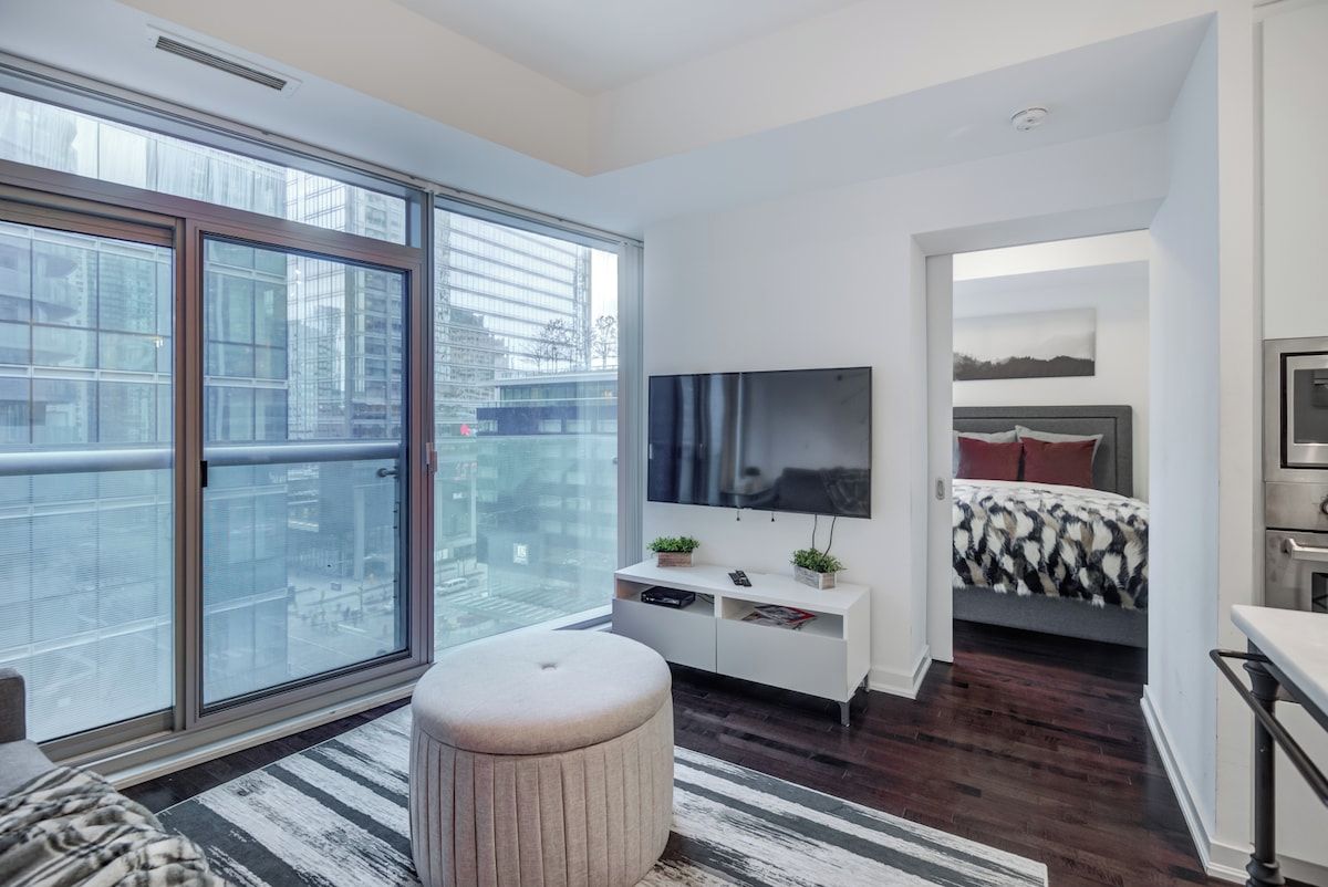 Een moderne slaapkamer met een groot raam, een flatscreen televisie, een bed met beddengoed met een patroon en een witte console tafel met planten.