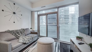 A modern living room with a sofa, ottoman, and a large window with a view of buildings. - Stylish 1Bed Den Unit in the heart of Dt (Toronto)